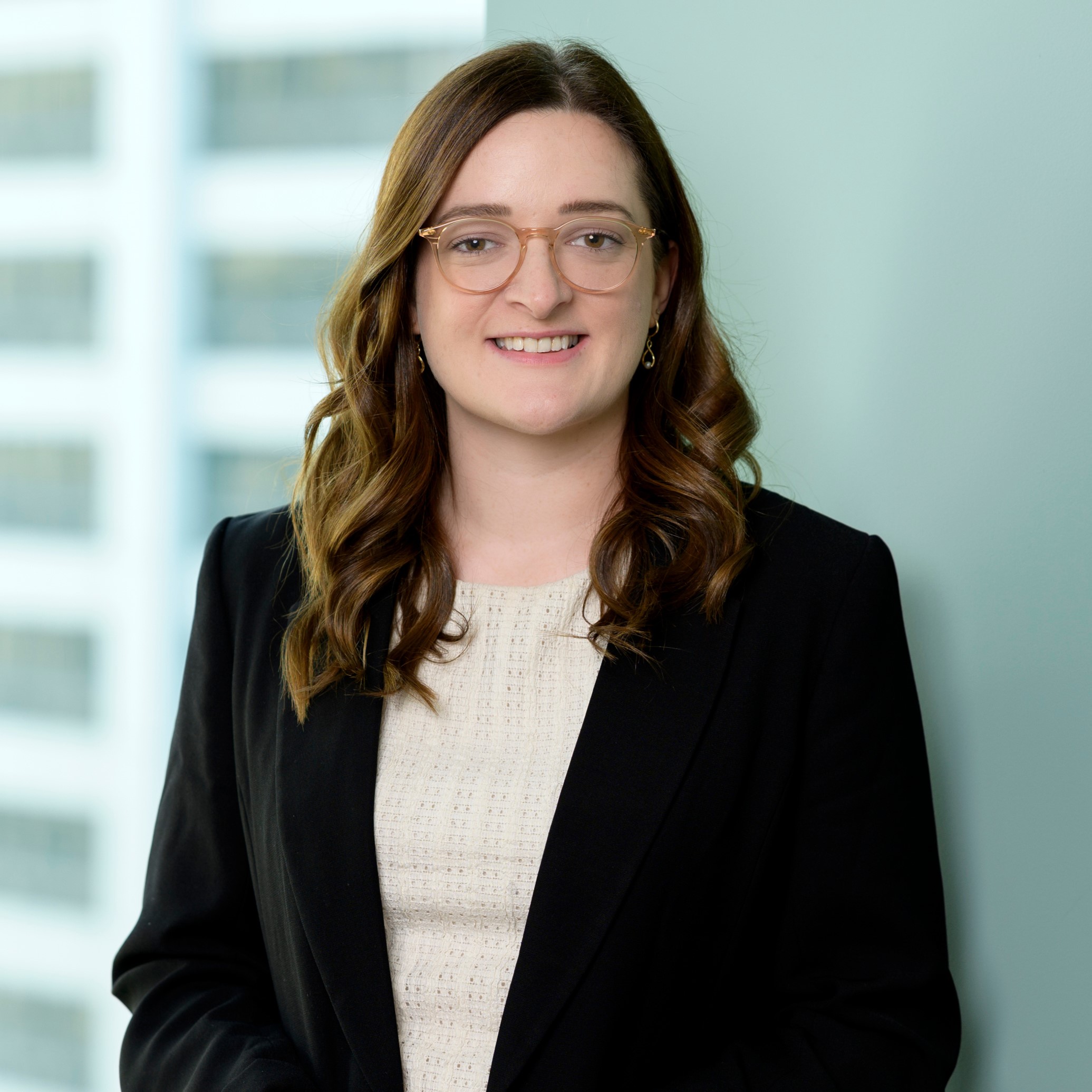 A woman with long brown hair, glasses, and a black blazer over a light-colored top smiles while standing indoors, with blurred windows and a light blue wall in the background.