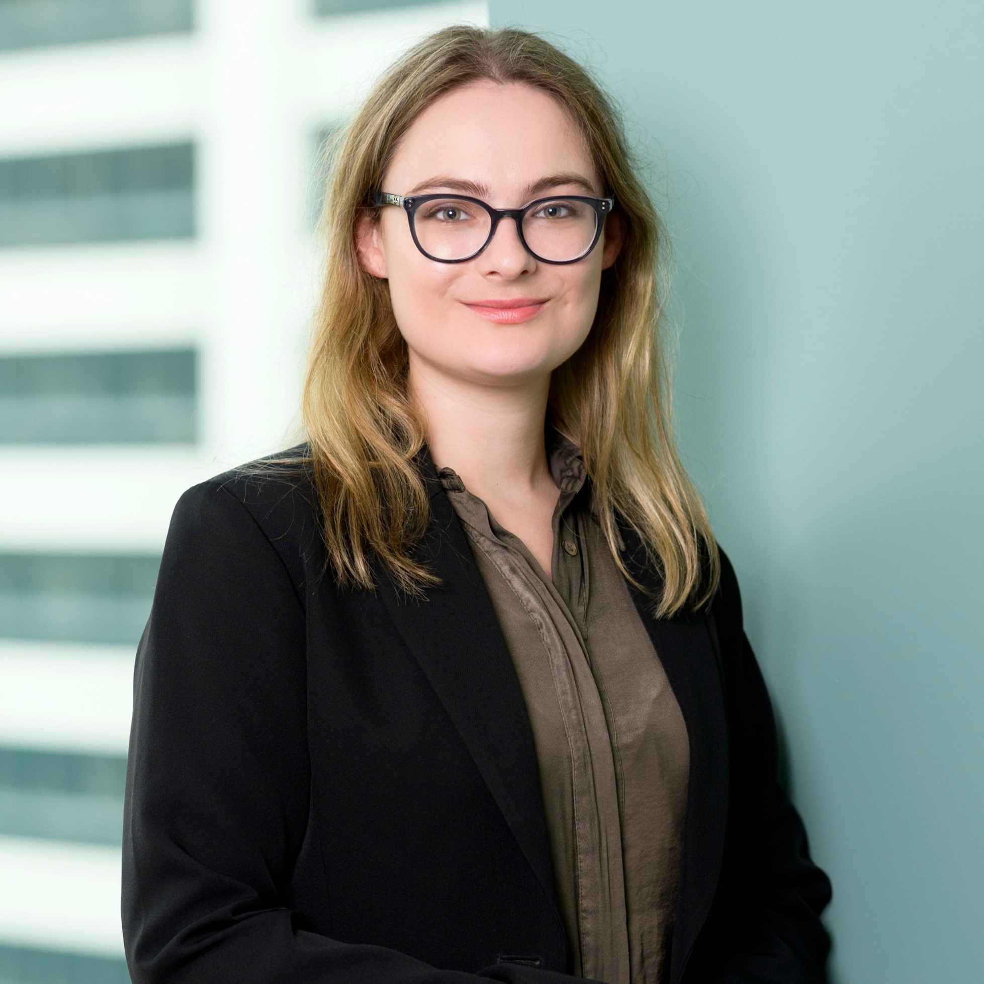 A woman with long blonde hair and glasses, wearing a dark blazer over a brown blouse, stands in front of a light green wall with blurred windows in the background, smiling slightly at the camera.