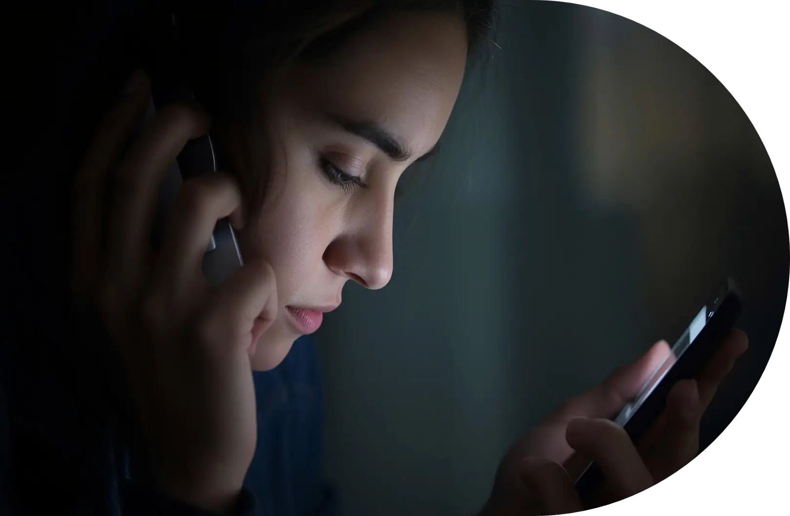 A person holds a phone to their ear while looking intently at another smartphone in their hand, illuminated by the screen's light in a dark environment.