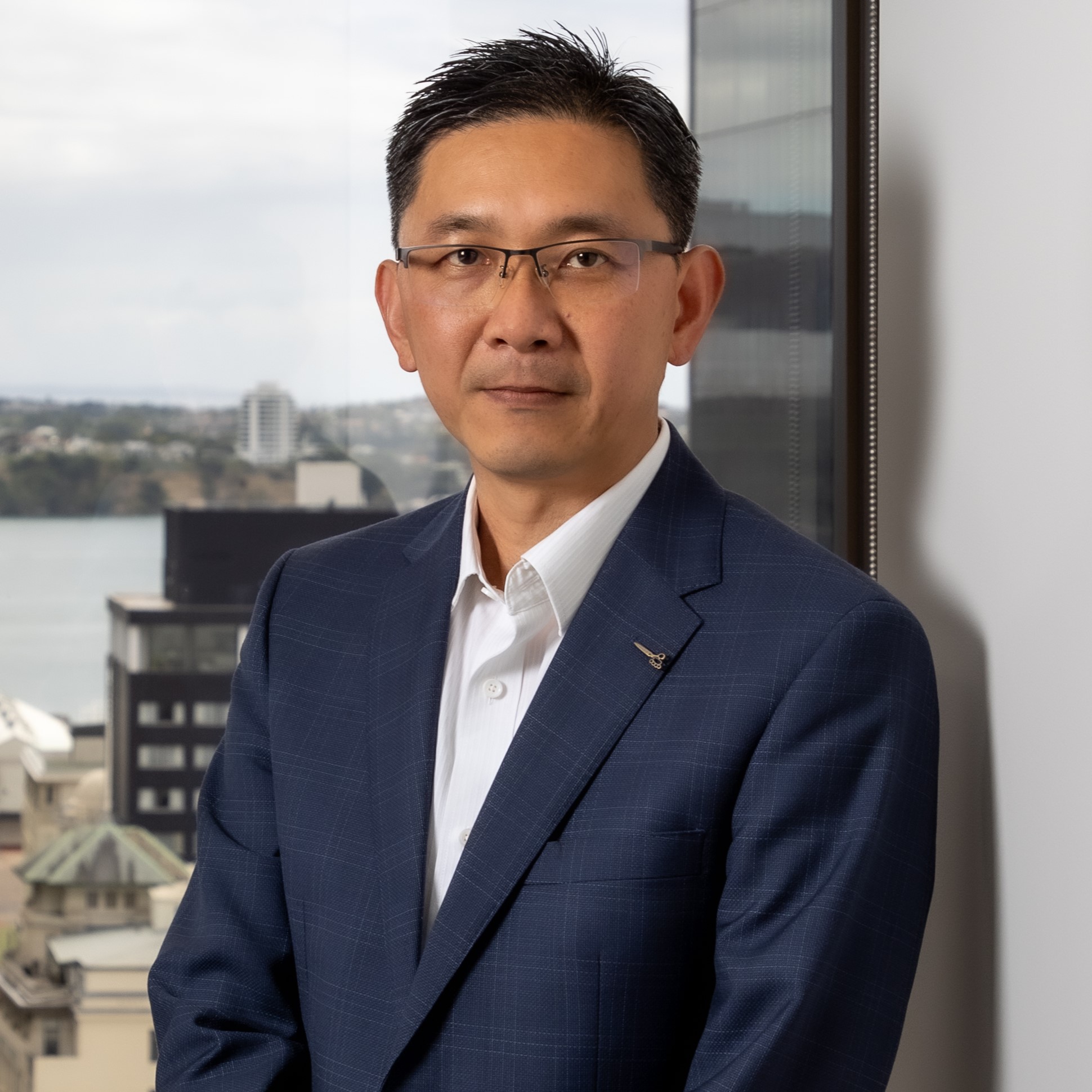 A man in a blue suit and glasses stands indoors by a window, with a cityscape and water visible in the background. He looks directly at the camera with a neutral expression.