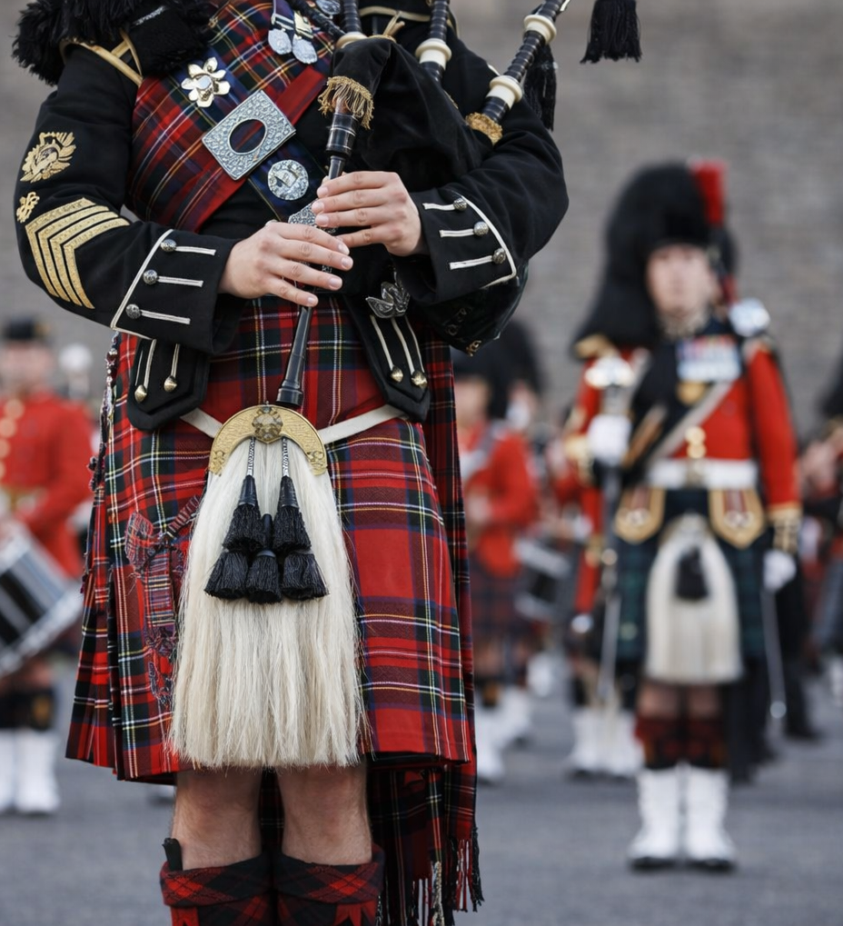 A bagpiper in traditional Scottish attire, including a red tartan kilt and sporran, plays the bagpipes during a parade, celebrating Scotland's cultural heritage as other uniformed musicians are blurred in the background.