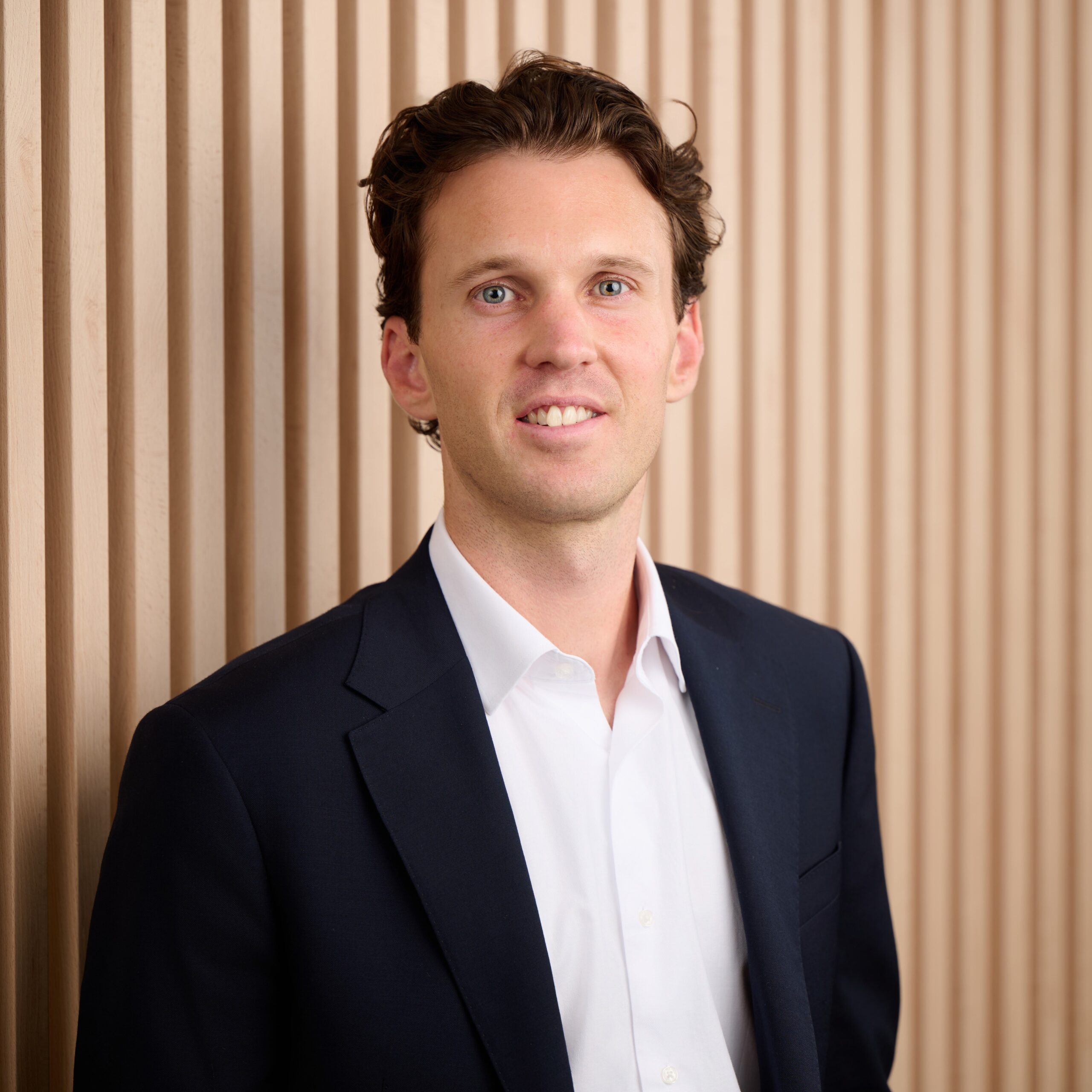 Connor Gavin, a man with wavy brown hair, wearing a dark suit jacket and white shirt, stands in front of a wall with vertical wooden slats, smiling slightly at the camera.