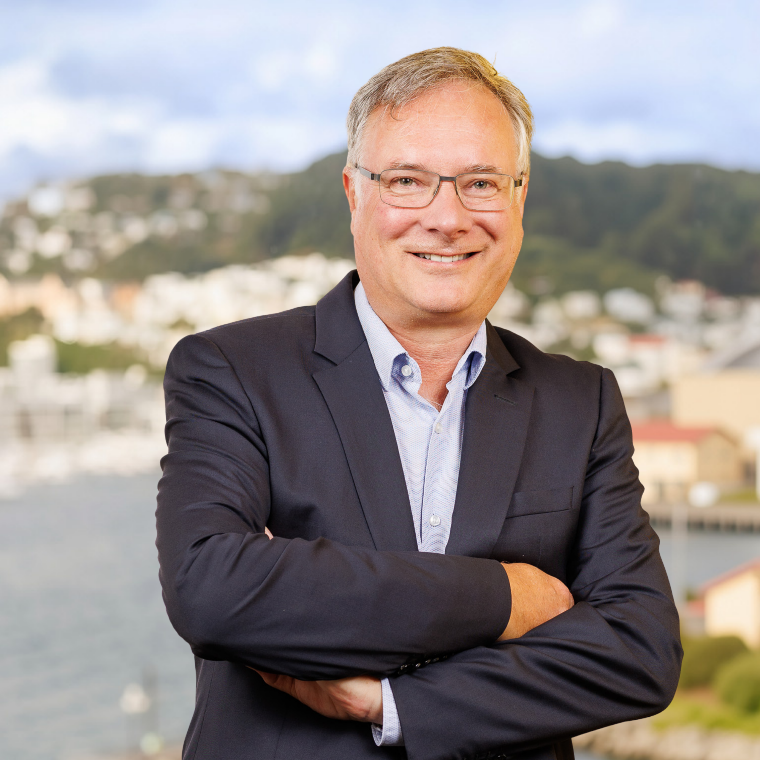 A middle-aged man in a suit and glasses stands outdoors, arms folded and smiling, with a blurred mix of city buildings and greenery behind him.