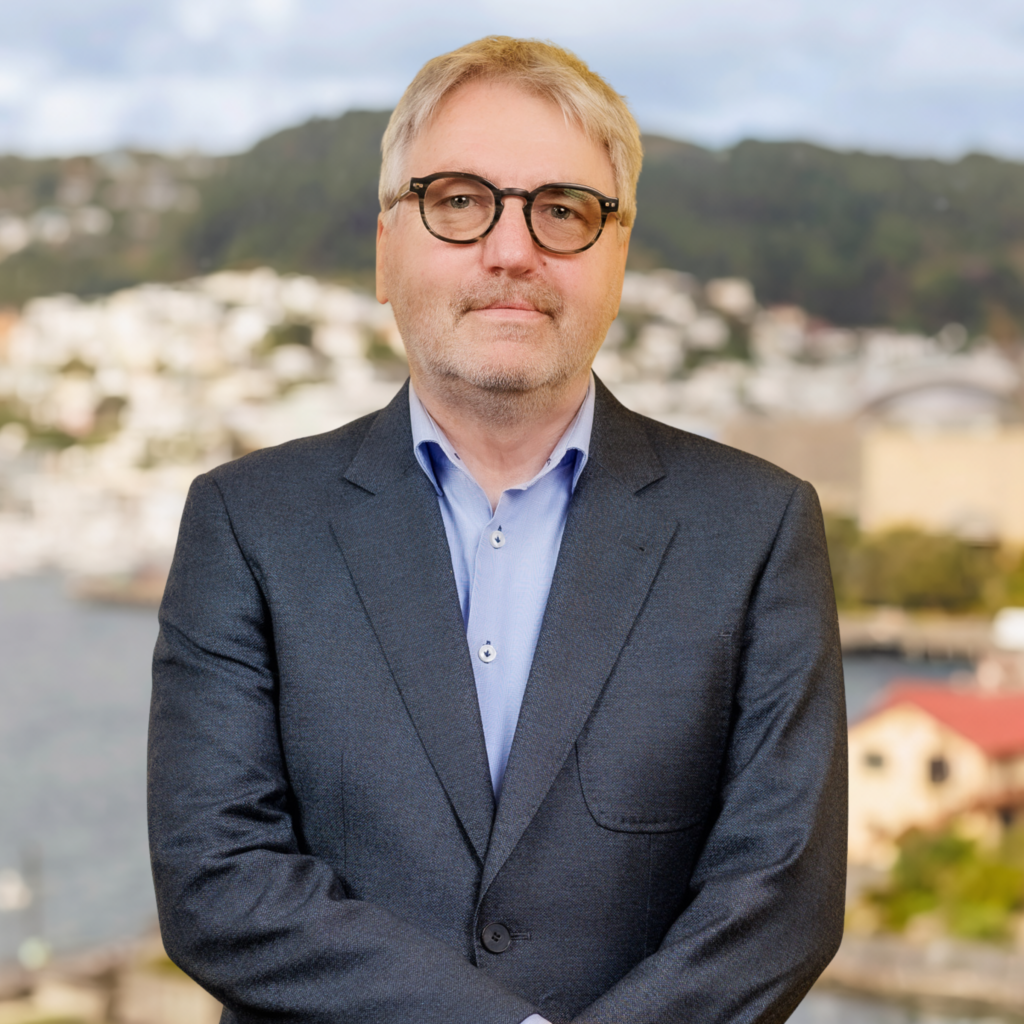 A man with grey hair and glasses, dressed in a dark suit and light blue shirt, stands outdoors in front of a blurred backdrop featuring water, buildings, and green hills.