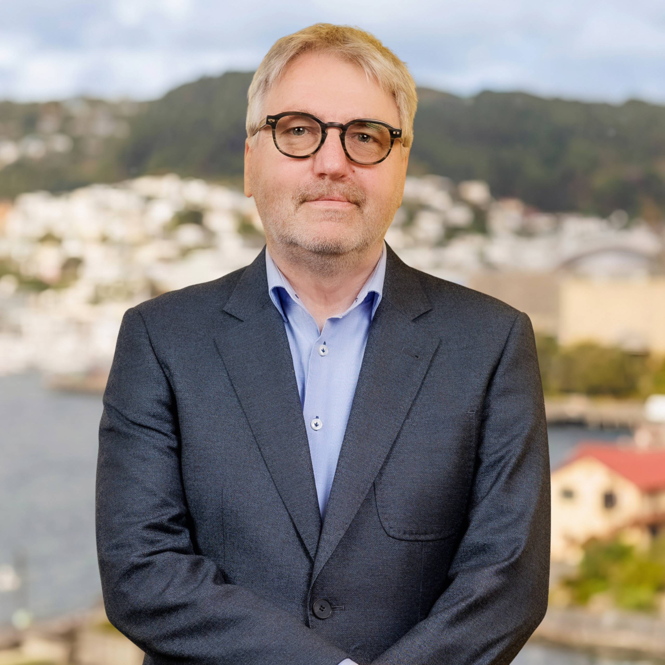 A man with grey hair and glasses, dressed in a dark suit and light blue shirt, stands outdoors in front of a blurred backdrop featuring water, buildings, and green hills.