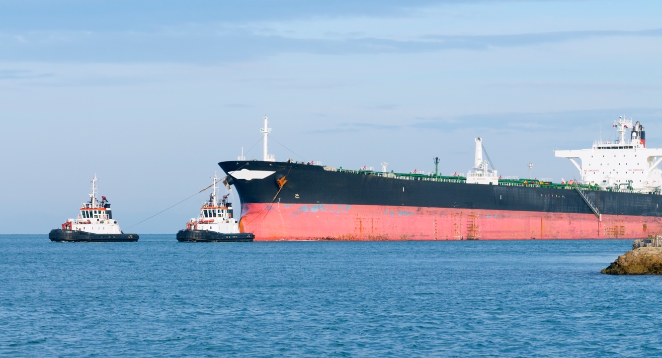 Two small tugboats, skilfully performing single post manoeuvres, escort a large black and red cargo ship across tranquil blue waters beneath a clear sky, while in the foreground part of a rocky shore with scattered tombstones is visible.