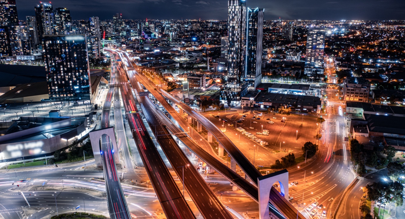 Aerial view of a city at night featuring glowing skyscrapers, bustling motorways, and streaks of car headlamps, highlighting the lively urban atmosphere and concentrated traffic flows. The scene encapsulates the energetic pulse of metropolitan life amid towering glass and steel structures.