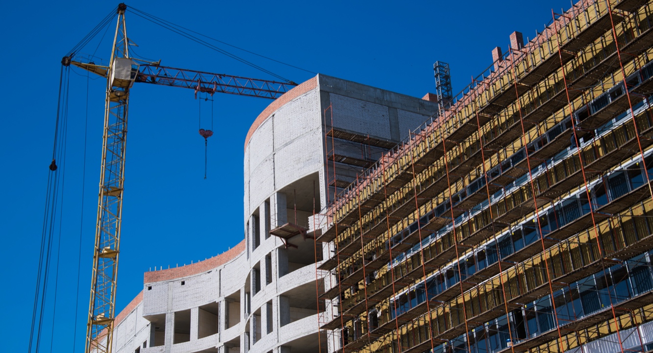 A yellow crane stands next to a modern building under construction, featuring scaffolding and exposed brick walls, highlighting construction expertise beneath a clear blue sky.