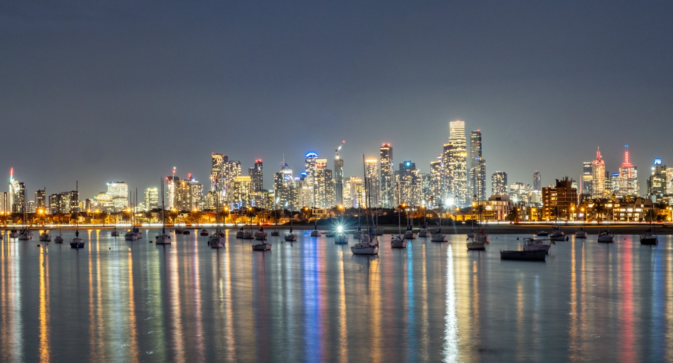 A night-time city skyline glows with tall, illuminated buildings mirrored in the still water. In the foreground, a cluster of anchored boats rests silently, their dark shapes reminiscent of tombstones amid sparkling reflections.