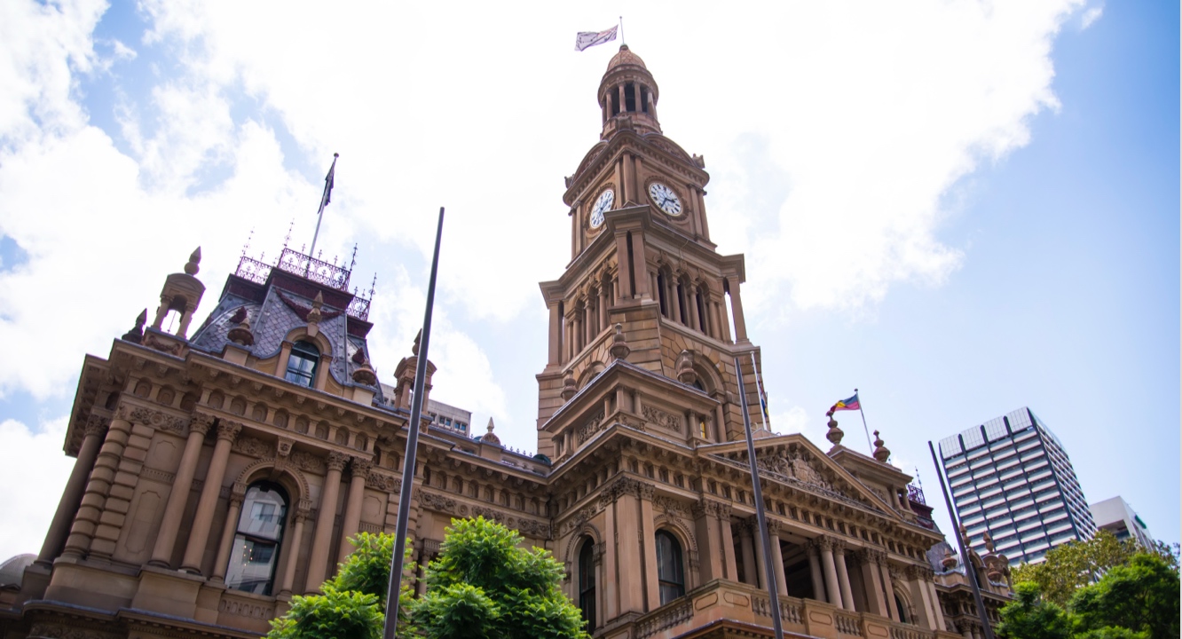 Historic sandstone building with a central clock tower, arched windows, flagpoles, and lush greenery in front, all set against a blue sky dotted with clouds. Behind the historic structure, modern high-rise buildings rise up, highlighting the blend of classic architectural craftmanship and contemporary urban development.