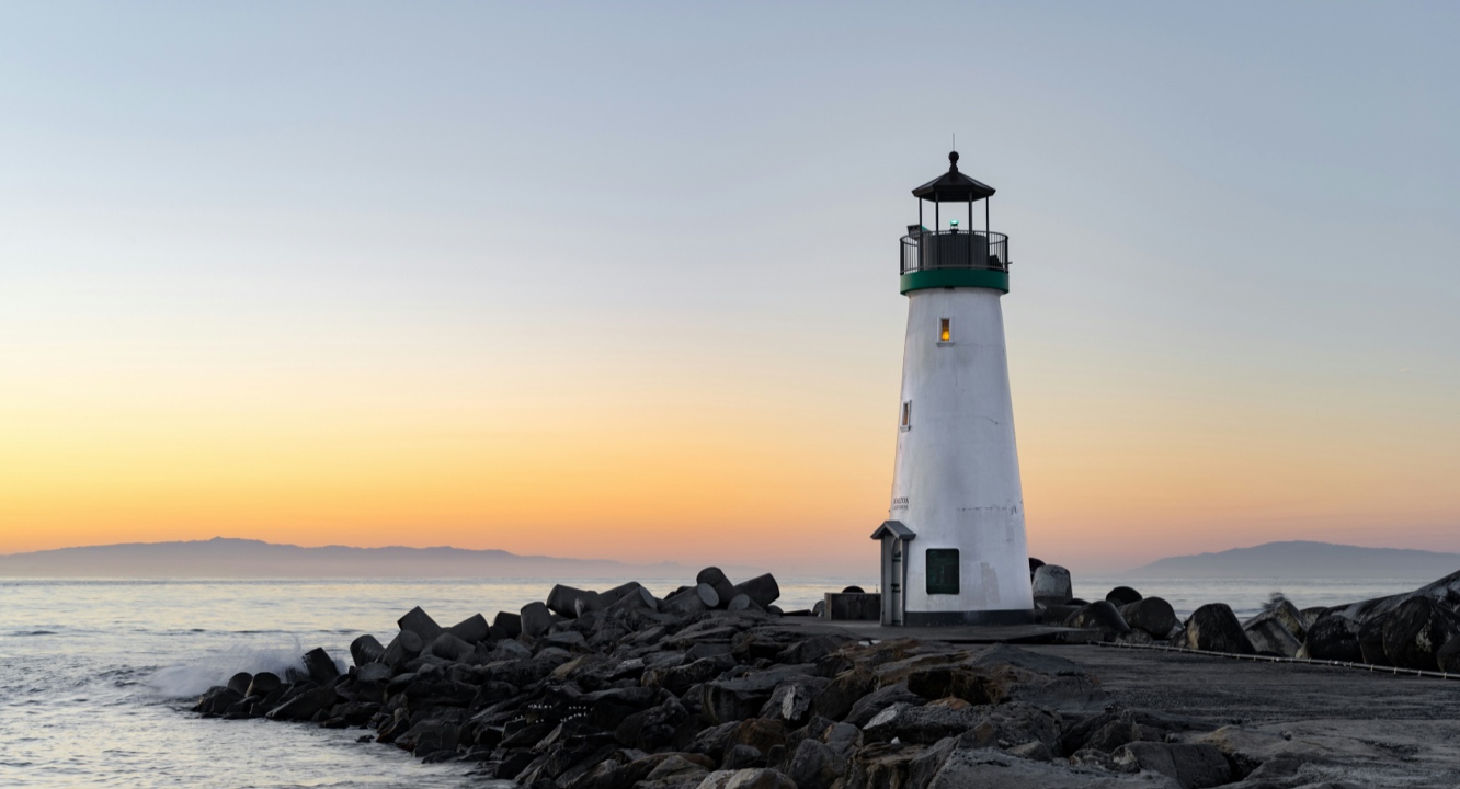 A white lighthouse with a green top stands on a rocky breakwater at sunset by the sea; serene waters reflect distant mountain silhouettes in the background, whilst weathered tombstones scattered nearby evoke tales of maritime history and lost seafaring knowledge.