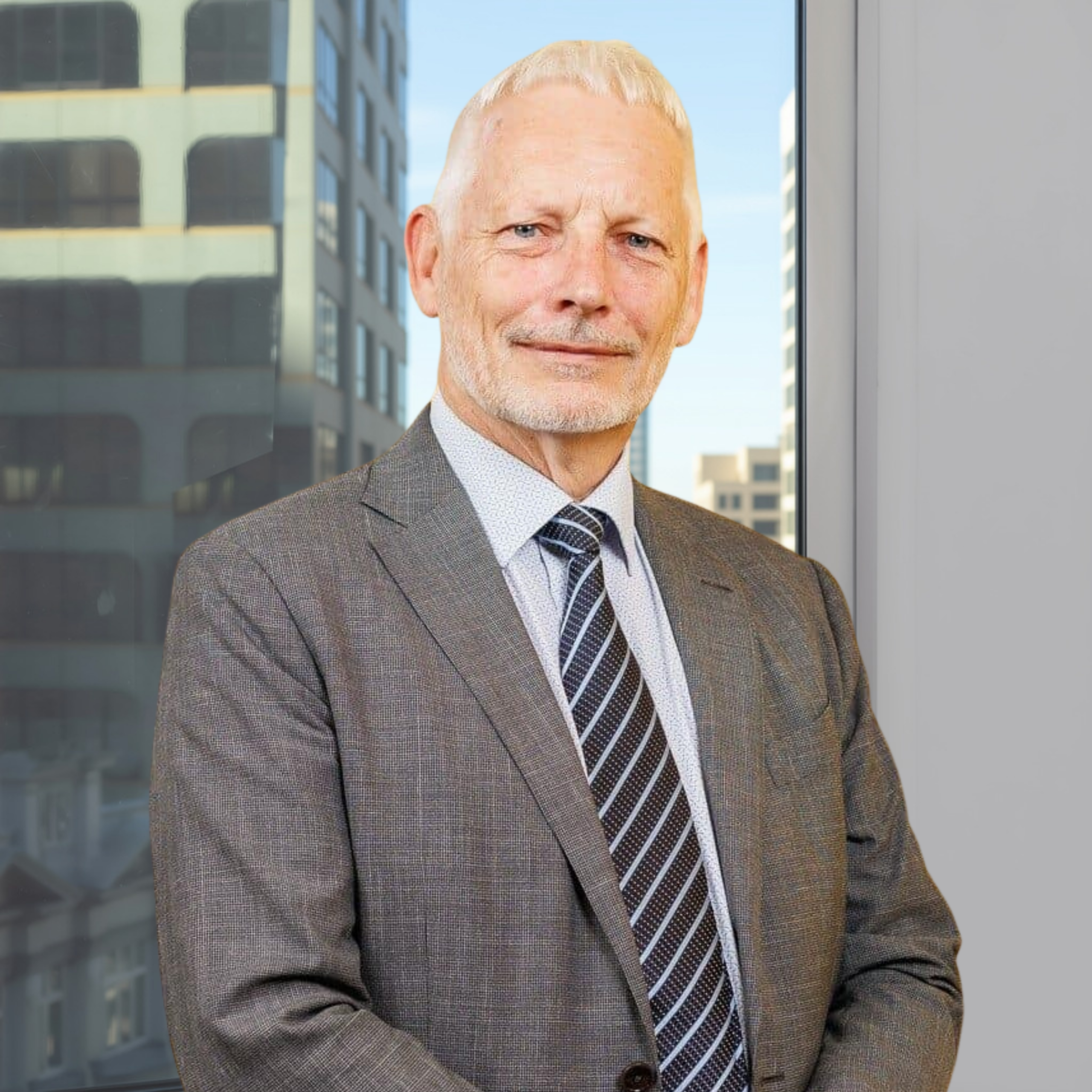 Older man with short white hair and a neatly trimmed beard, dressed in a grey suit, blue shirt, and a striped tie, standing before a large window overlooking city buildings.
