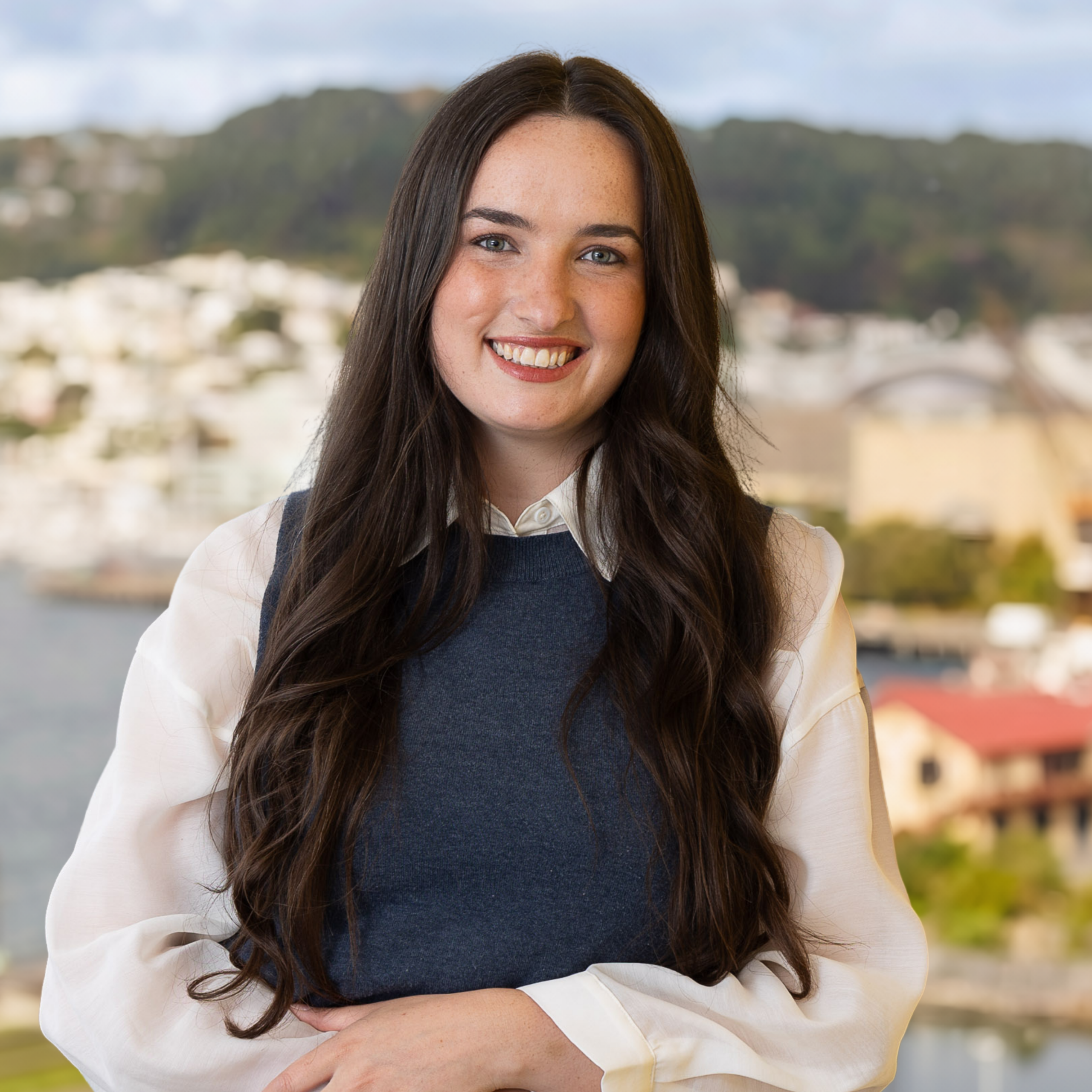 A young woman with long brown hair, dressed in a white blouse and blue waistcoat, smiles whilst standing outside against a backdrop of blurred buildings, water, and green hills.