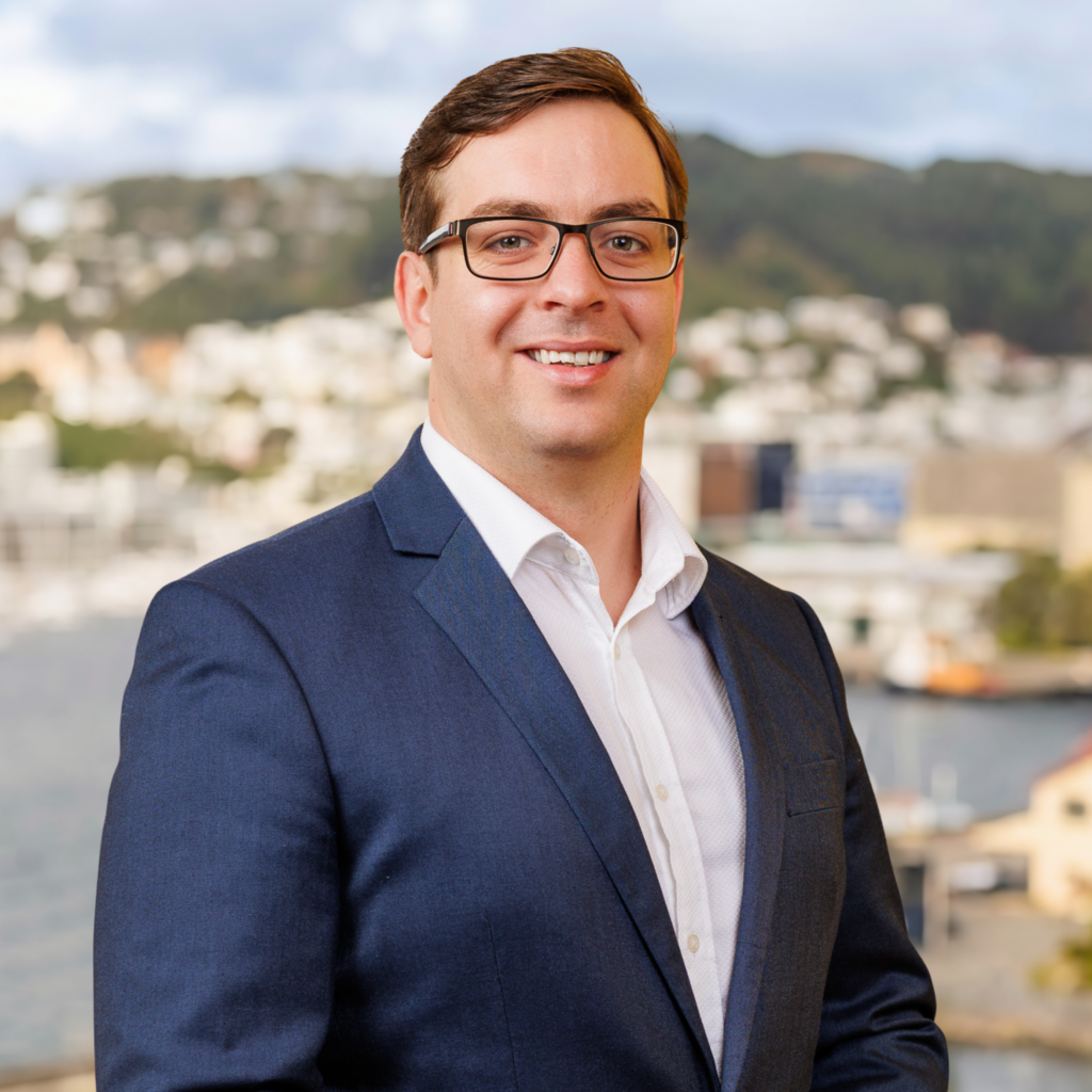 A man in a navy suit and glasses smiles whilst standing outdoors, with a blurred cityscape and hills in the background.