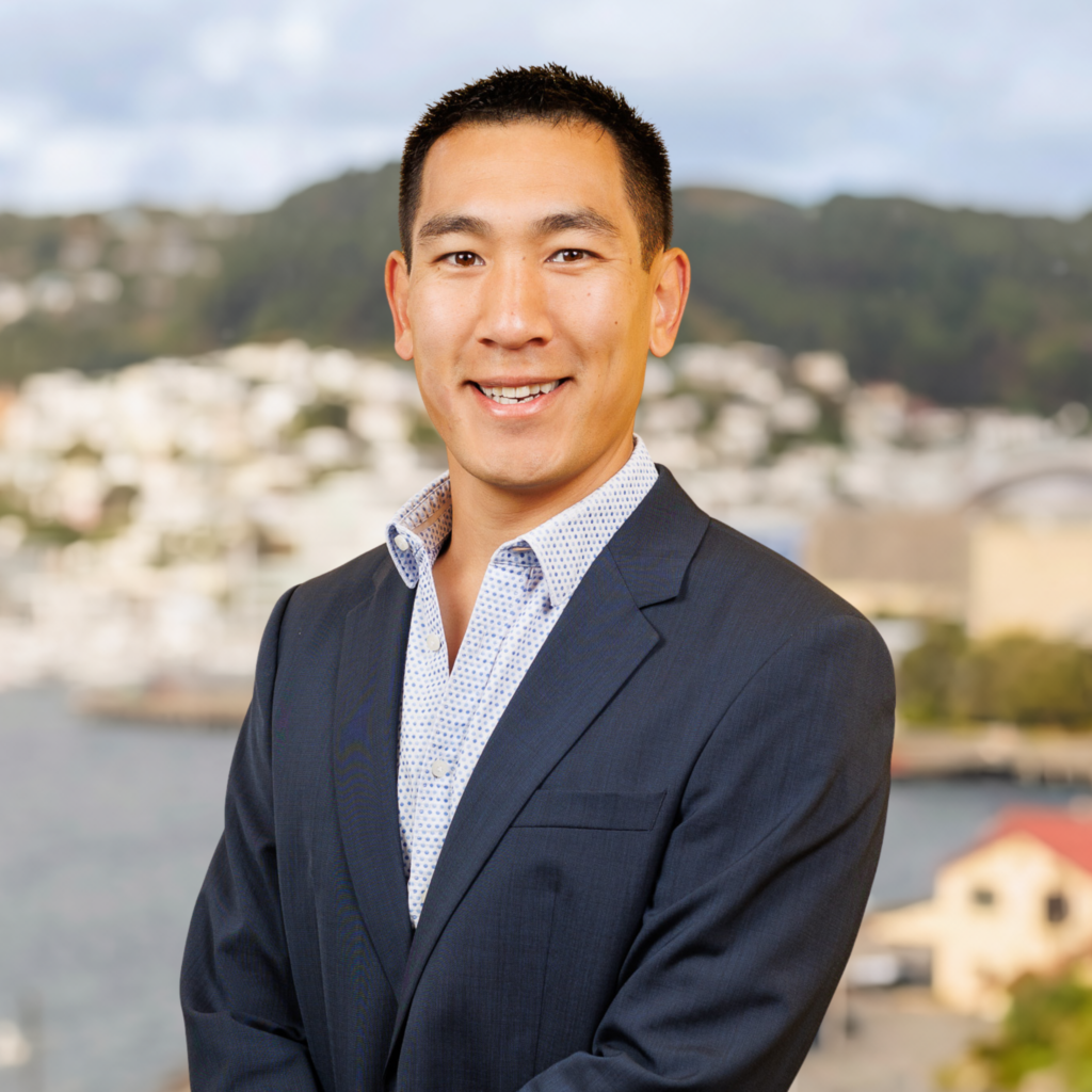 A man in a navy suit and patterned shirt stands outside, smiling towards the camera. Behind him, the out-of-focus background reveals hills, buildings, and water, indicating he is in a coastal city environment.