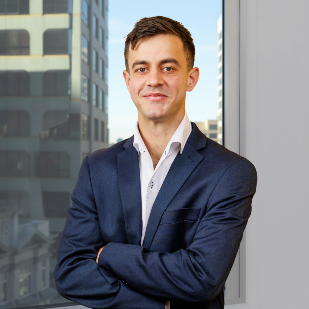 A man in a navy suit stands with arms crossed, smiling in an office with large windows overlooking city buildings.