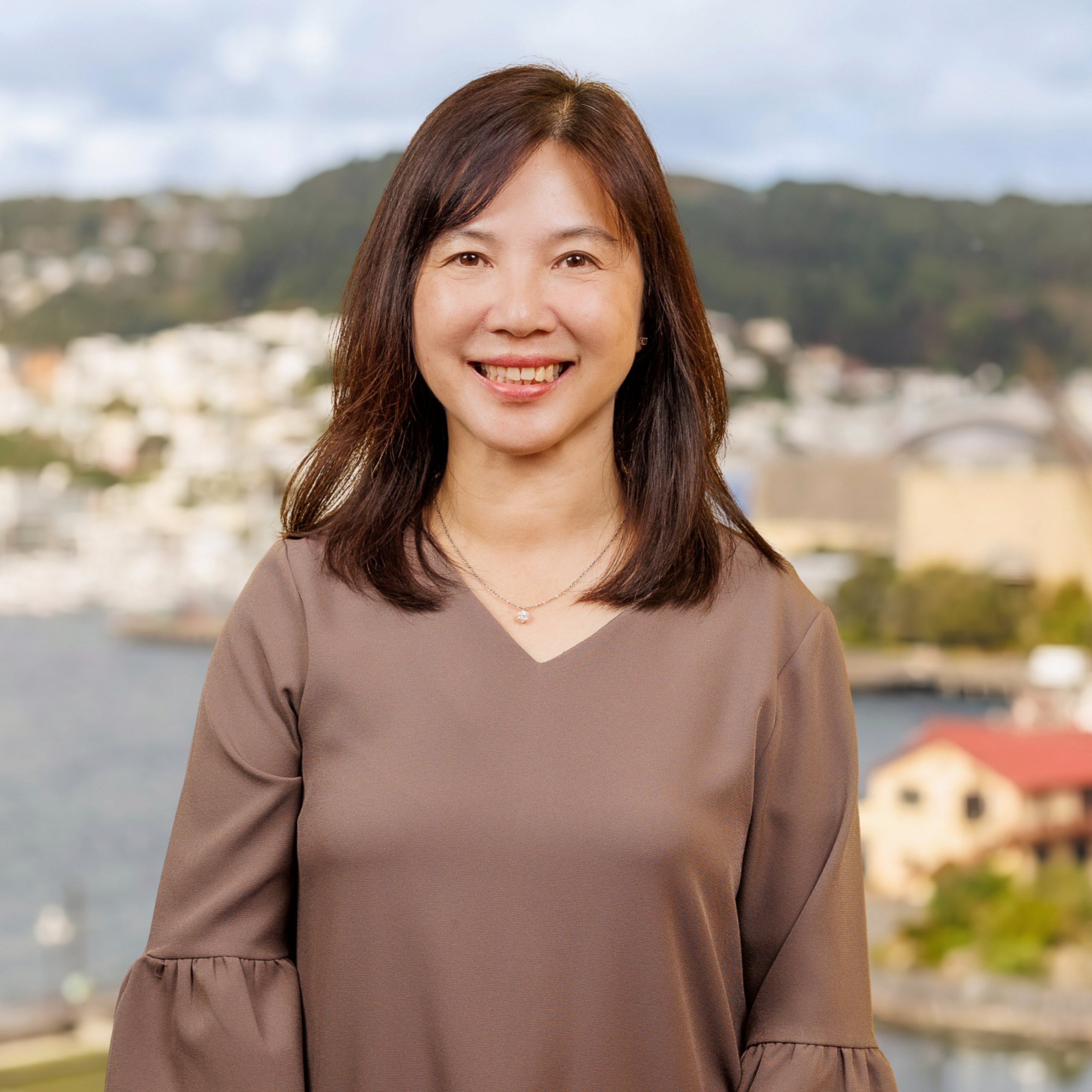 A woman with shoulder-length brown hair smiles at the camera, standing outdoors with a blurred background of buildings, water, and hills under a partly cloudy sky.