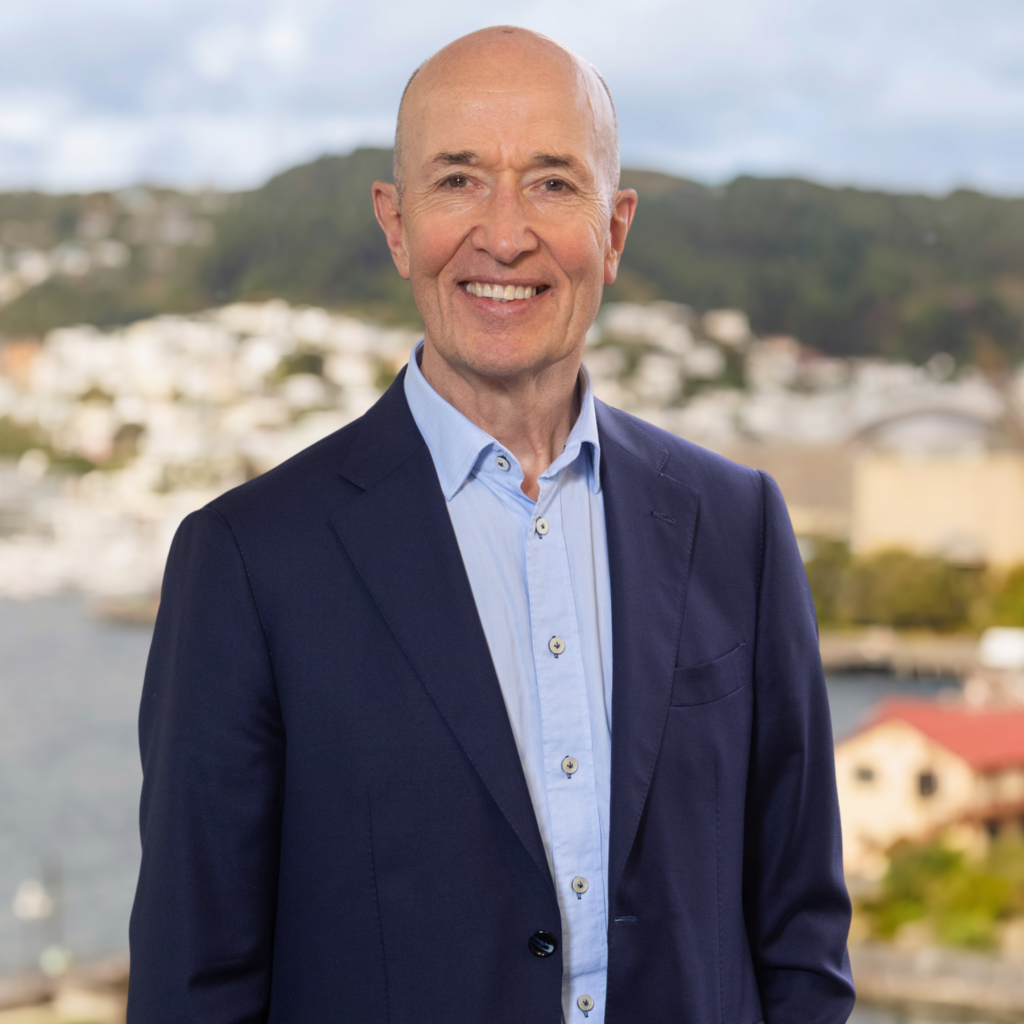 Bald man in a blue suit and light blue shirt smiles outdoors with hills, water, trees, and buildings visible in the scenic background beneath a partly cloudy sky.