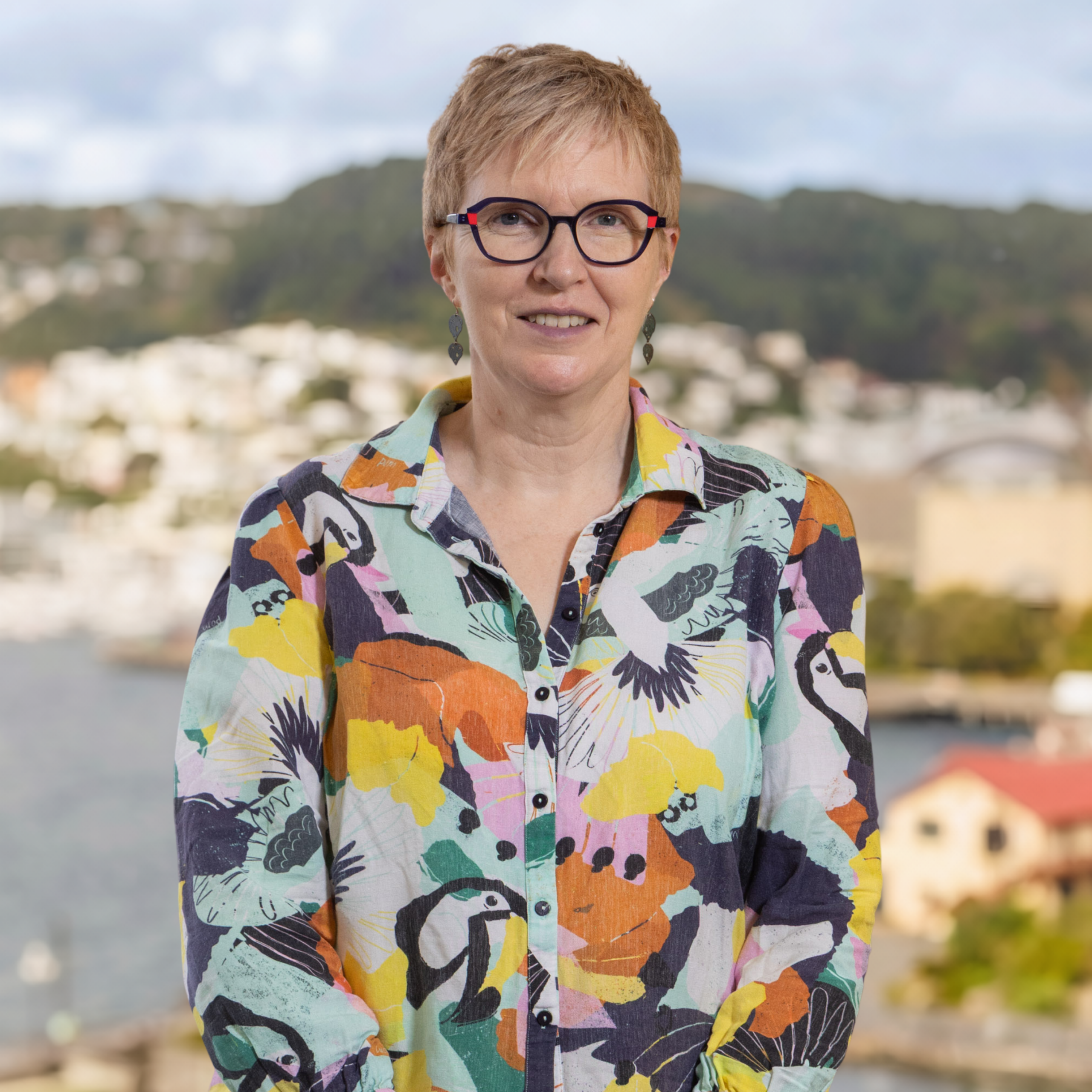 A person with short hair and glasses stands outside in a vibrant bird-patterned shirt, set against a softly blurred backdrop of water, trees, and distant buildings.