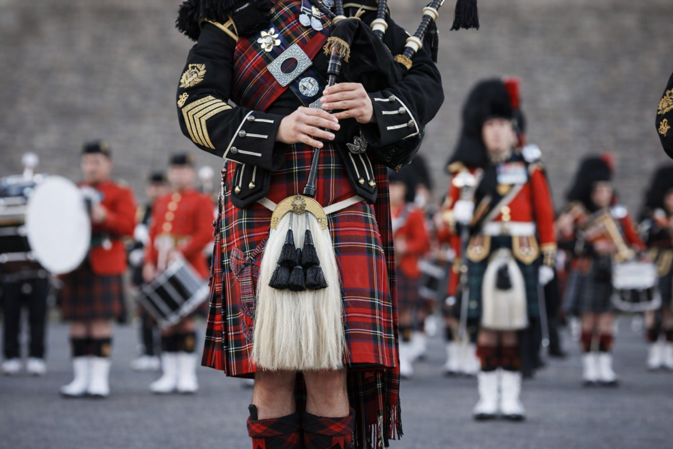 A bagpiper dressed in traditional Scottish clothing performs, accompanied by other similarly attired musicians standing behind him at a parade or ceremony, exemplifying the role of cultural heritage in driving growth and supporting regional development.