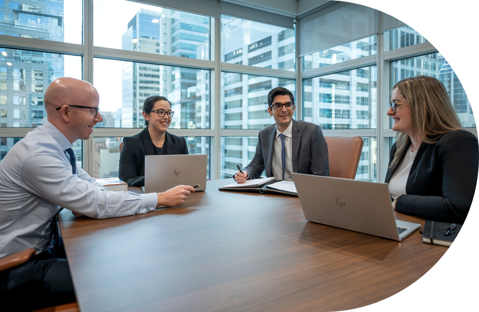 Four professionals sit around a conference table with laptops, engaged in a meeting in a modern office featuring large windows that reveal city buildings outside.
