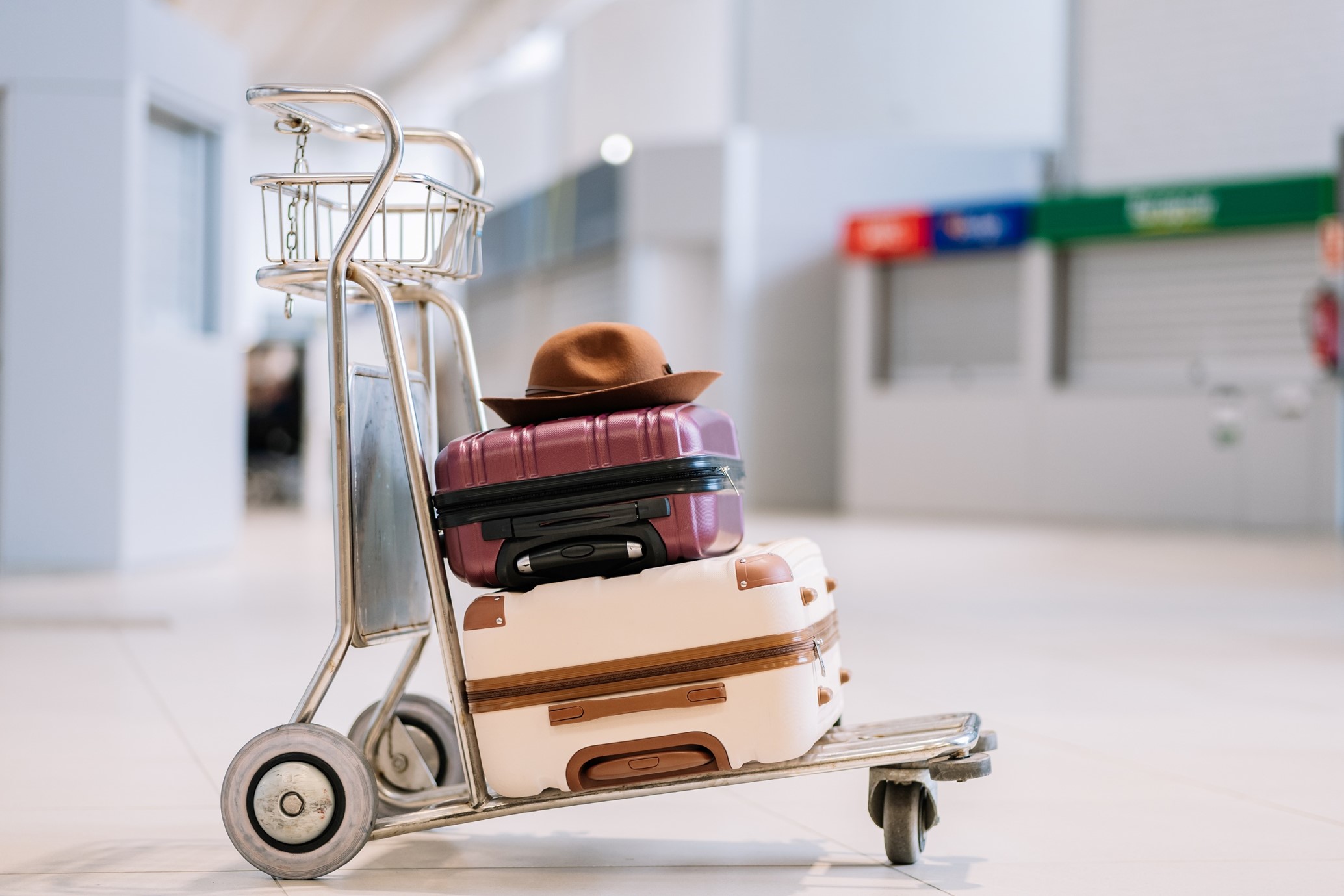 A luggage trolley with a brown hat resting on top, as well as a purple suitcase and a white suitcase, stands alone in an empty airport terminal, subtly representing the economic effects of tourism and emphasising the significance of travel within global tourism economics.